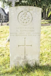 Colour photograph of a white gravestone on a sunny day. Longer grass is growing around the bottom of the stone.